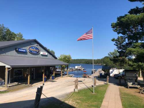 A lakeside marina with a flag, boats docked, and a building labeled "Hadberg's Marina" under a clear blue sky.
