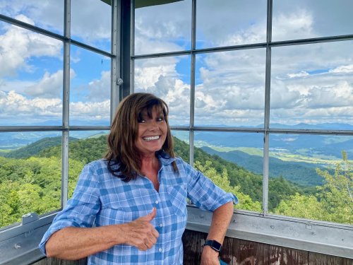 A woman in a blue checkered shirt smiles and gives a thumbs up inside a lookout tower with a scenic mountain view.