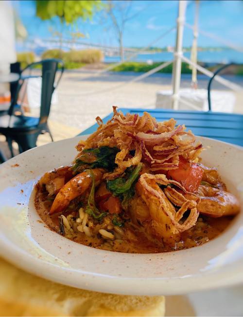 A bowl of shrimp and rice topped with crispy onions, set against a scenic outdoor backdrop by the water.