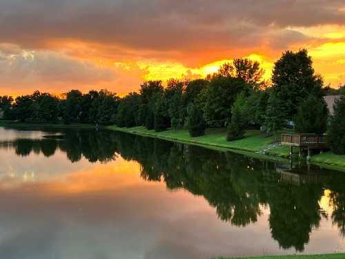 A serene lake at sunset, reflecting vibrant orange and yellow hues, surrounded by lush green trees.