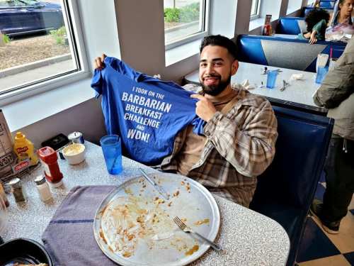 A smiling man holds up a blue shirt that says "I took on the Barbarian Breakfast Challenge and won!" at a diner.