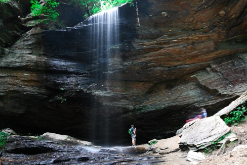 A person stands near a waterfall cascading down rocky cliffs in a lush, green forest setting.