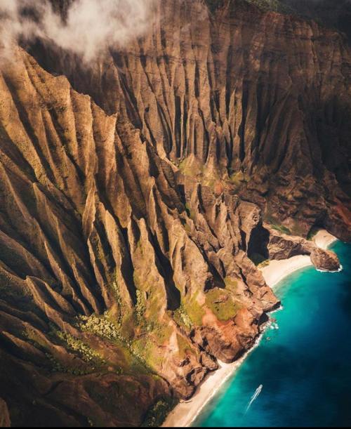Aerial view of rugged cliffs meeting a turquoise ocean, with a small boat near a sandy beach.
