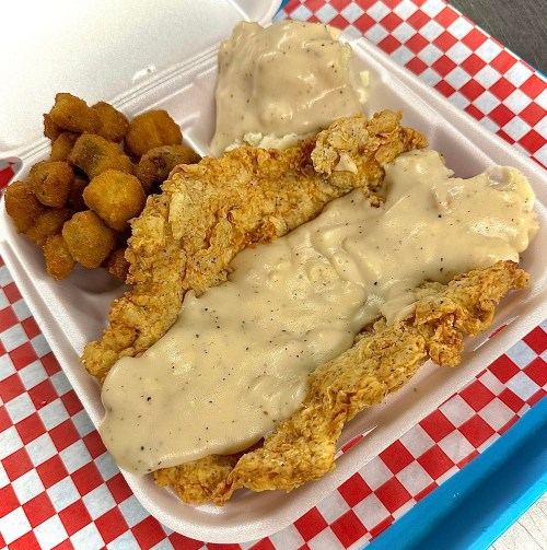 A takeout container with fried chicken strips covered in gravy, mashed potatoes, and a side of fried okra.