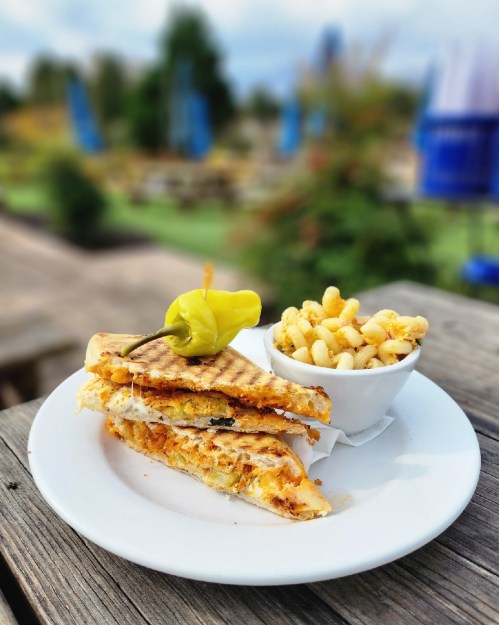 A grilled sandwich with melted cheese and vegetables, served with a side of macaroni and a pepper on top.