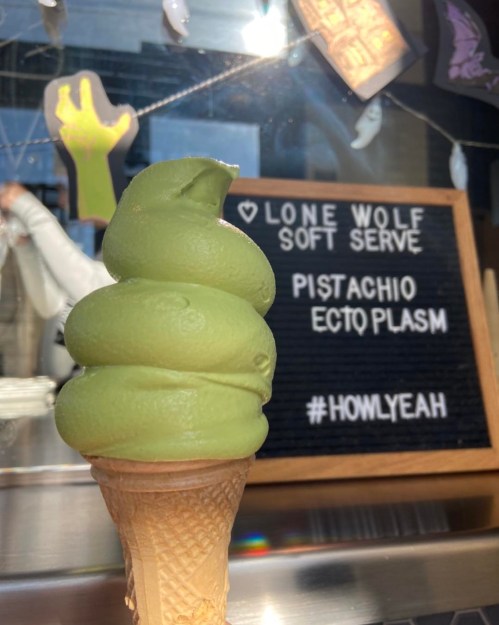 A green pistachio soft serve ice cream cone in front of a sign that reads "Lone Wolf Soft Serve" and "#HOWLYEAH."