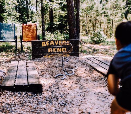 A sandy horseshoe pit with a sign reading "Beaver Bend" surrounded by trees and two chairs in the background.