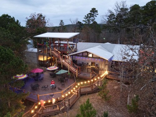 Aerial view of a rustic lodge with a deck, colorful umbrellas, and festive lights, surrounded by trees.