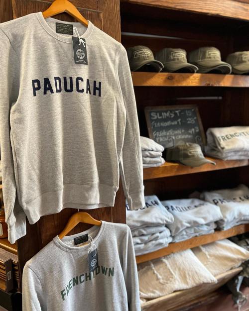 A gray sweatshirt with "PADUCAH" printed on it hangs on a wooden display, surrounded by other clothing items.