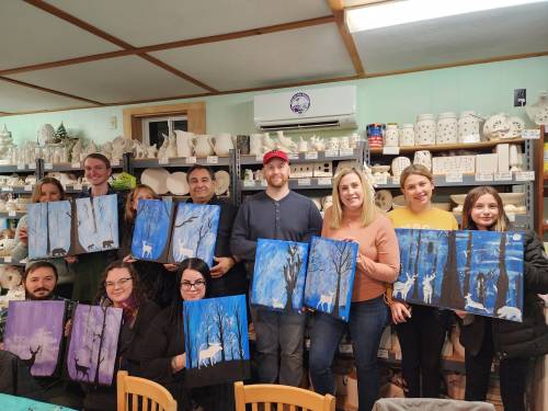 A group of people holding their painted canvases in a cozy art studio, showcasing winter scenes with trees and deer.