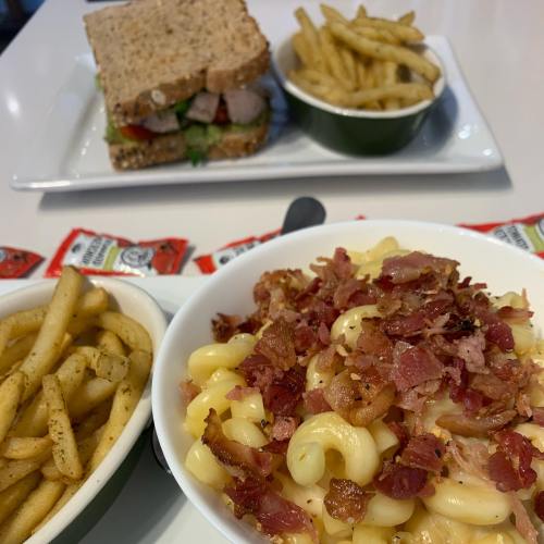 A bowl of macaroni and cheese topped with bacon, served with fries and a sandwich in the background.