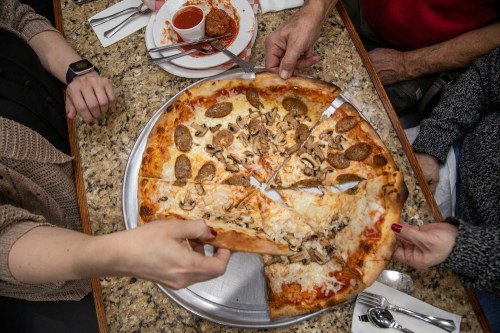 A group of people sharing a large pizza topped with sausage and mushrooms, with plates and sauce in the background.
