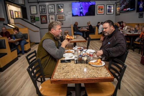 Two men sit at a table in a restaurant, eating pizza and chatting, with other patrons and a TV in the background.