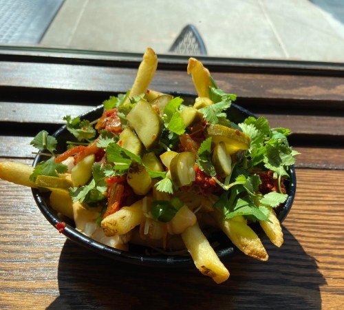 A bowl of fries topped with pickles, herbs, and a mix of vegetables, sitting on a wooden table.