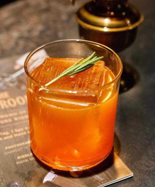 A cocktail in a glass with a large ice cube and a sprig of rosemary, resting on a coaster.