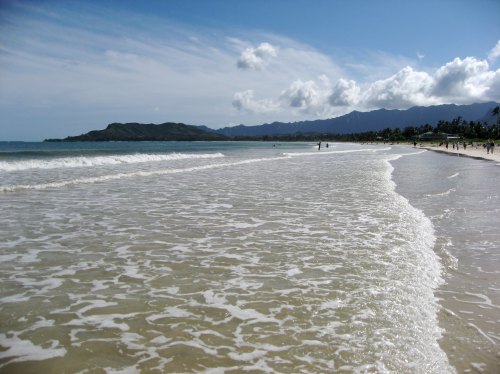 A serene beach scene with gentle waves, distant mountains, and a partly cloudy sky. People stroll along the shore.