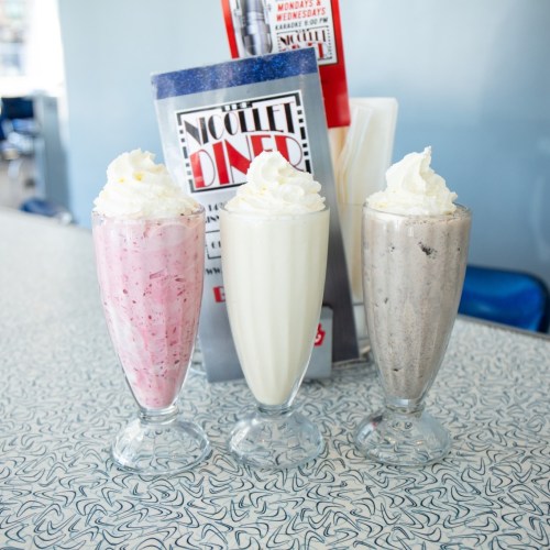 Three milkshakes in tall glasses: pink strawberry, white vanilla, and gray cookies and cream, topped with whipped cream.