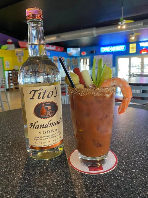 A bottle of Tito's Handmade Vodka next to a garnished Bloody Mary with shrimp, celery, and a salted rim on a table.