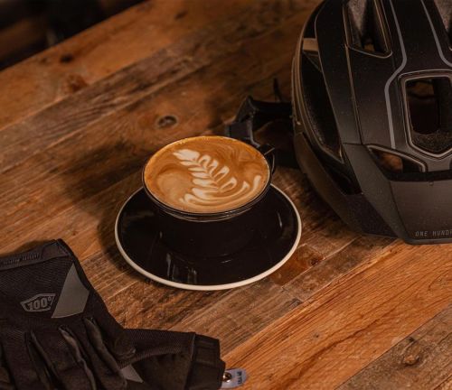 A coffee cup with latte art sits next to a black bike helmet and gloves on a wooden table.