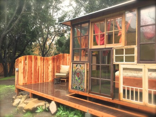 A cozy wooden cabin with large windows, colorful curtains, and a wooden deck surrounded by greenery.