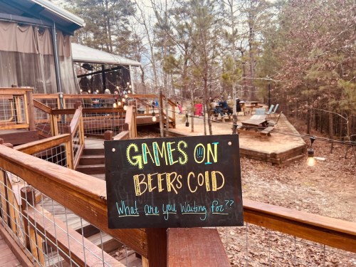 A wooden sign reads "GAMES ON BEERS COLD" in a rustic outdoor setting with trees and a gathering area in the background.