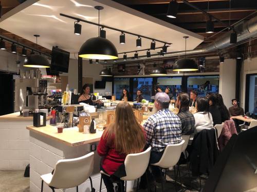 A group of people seated at a bar in a modern café, listening to a speaker at the front. Warm lighting and a cozy atmosphere.