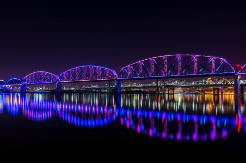 A brightly lit bridge with purple and blue lights reflecting on the water at night. City skyline in the background.