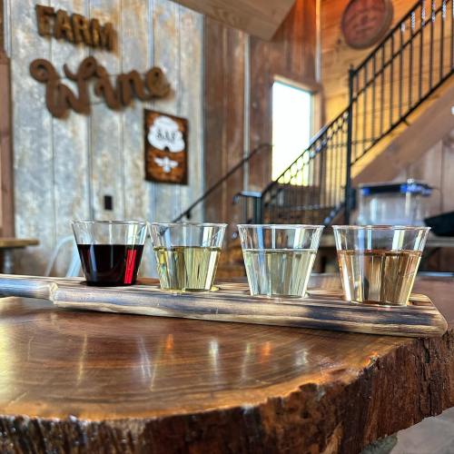 Four small glasses of wine on a wooden tray, set against a rustic interior with a "Farm Wine" sign.