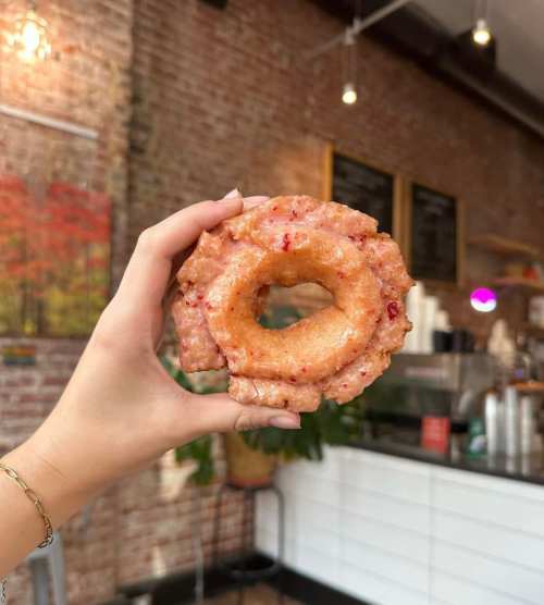 A hand holding a glazed strawberry donut in a cozy café setting with brick walls and a coffee counter in the background.