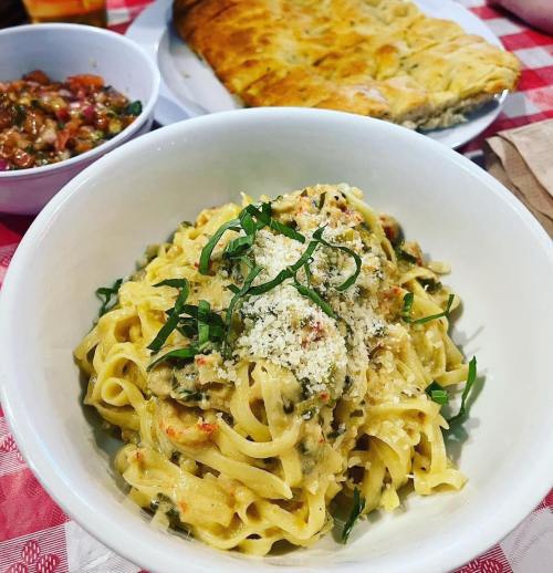 A bowl of creamy pasta topped with herbs and cheese, accompanied by a side of bread and salsa on a checkered tablecloth.