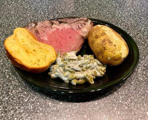 A plate with sliced roast beef, a baked potato, green bean casserole, and two pieces of garlic bread.