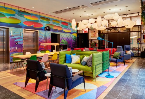 A vibrant lounge area with colorful furniture, large light fixtures, and a mural, featuring seating and tables for socializing.