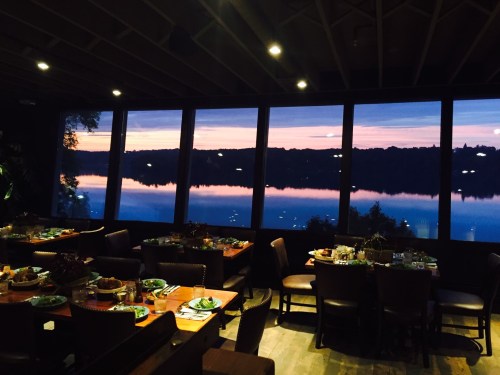 A dining area with tables set for a meal, featuring large windows showcasing a sunset over a calm lake.