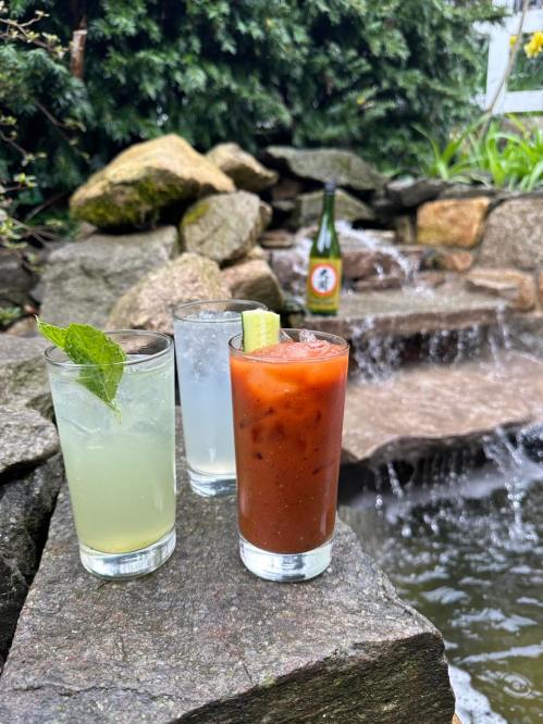 Three cocktails on a stone ledge by a water feature: a mint drink, a clear cocktail, and a Bloody Mary.