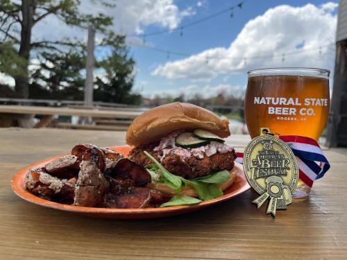 A crispy chicken sandwich with pickles and slaw, served with fries and a beer in a glass, outdoors with trees in the background.