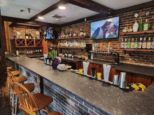 A rustic bar with a stone countertop, shelves of liquor, and a TV, featuring wooden stools and decorative flowers.