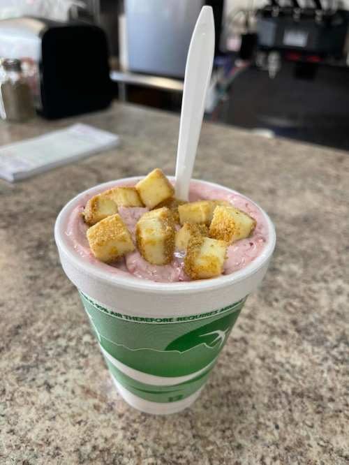 A cup of pink smoothie topped with small cubes of fruit, placed on a countertop. A white spoon is inside the cup.