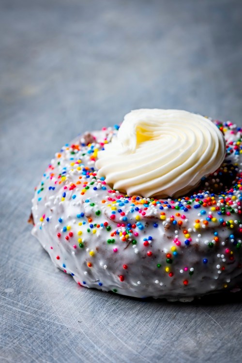 A frosted donut topped with colorful sprinkles and a swirl of cream on a textured gray surface.