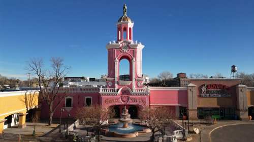A colorful pink building with a fountain, featuring a statue on top, surrounded by trees and shops under a clear blue sky.
