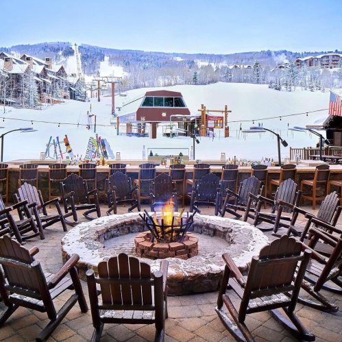 A cozy outdoor seating area with a fire pit, overlooking a snowy ski resort and chairlift in the background.