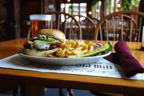A plate with a burger topped with cheese and bacon, fries, and a glass of beer on a wooden table in a cozy setting.
