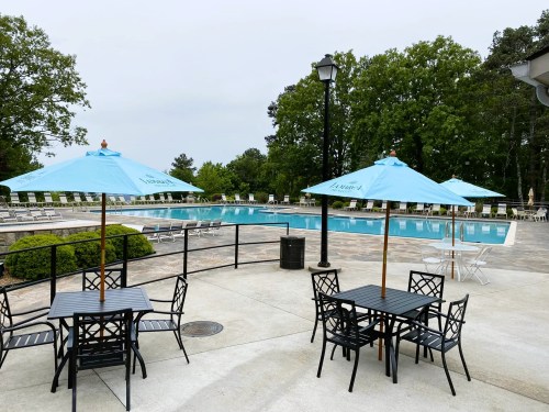 A serene pool area with blue umbrellas, tables, and lounge chairs surrounded by greenery on a cloudy day.