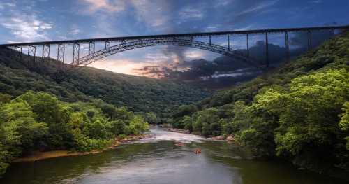 A scenic view of a river surrounded by lush greenery, with a large bridge arching over it under a dramatic sky.