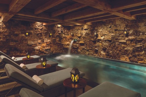 A serene spa interior with a stone wall, a tranquil pool, and cozy lanterns illuminating lounge chairs.