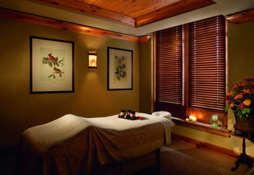 A serene spa room with a massage table, candles, and artwork, featuring warm lighting and wooden accents.