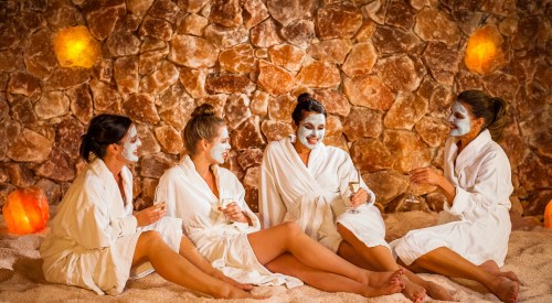 Four women in white robes enjoy a spa day, sitting on salt stones with face masks and drinks, surrounded by glowing lamps.