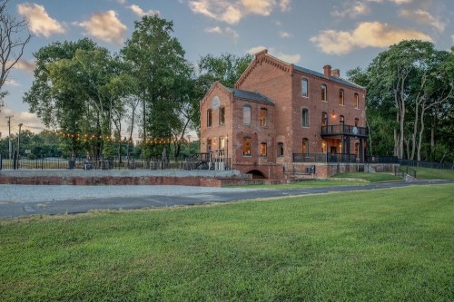 Historic brick building with large windows, surrounded by greenery and string lights, set against a twilight sky.