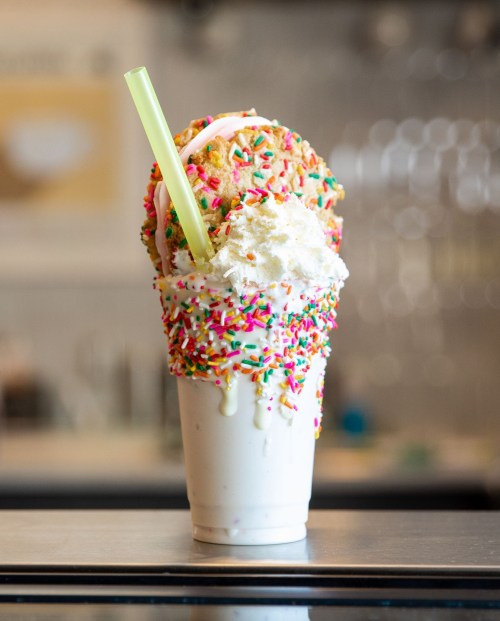 A tall milkshake topped with whipped cream, colorful sprinkles, and a cookie, served with a green straw.
