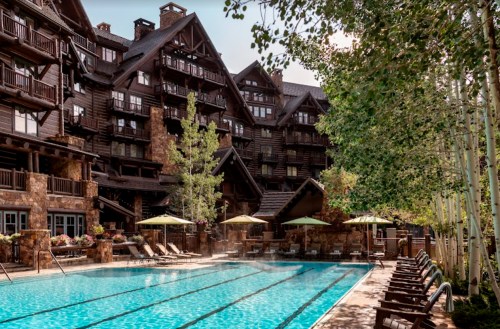 A luxurious hotel with a pool surrounded by trees and lounge chairs, featuring rustic architecture and a serene atmosphere.