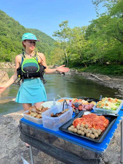 A person in a blue outfit stands by a river, showcasing a picnic spread with various foods on a table.